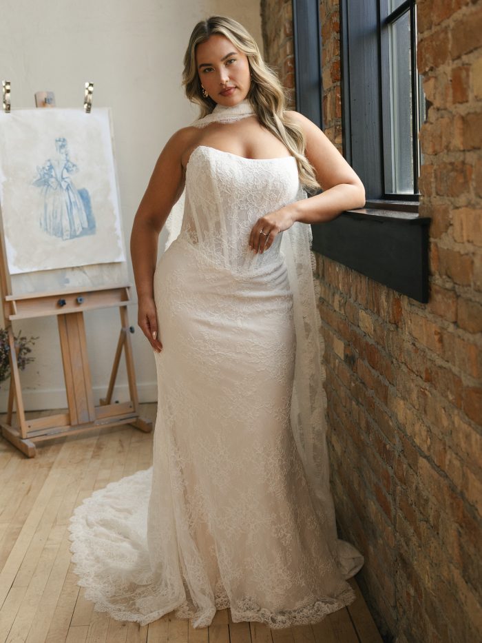 Model wearing Ariadne by Maggie Sottero, a strapless Chantilly lace sheath wedding dress with exposed boning bodice, cat eye neckline, and extended illusion lace train.