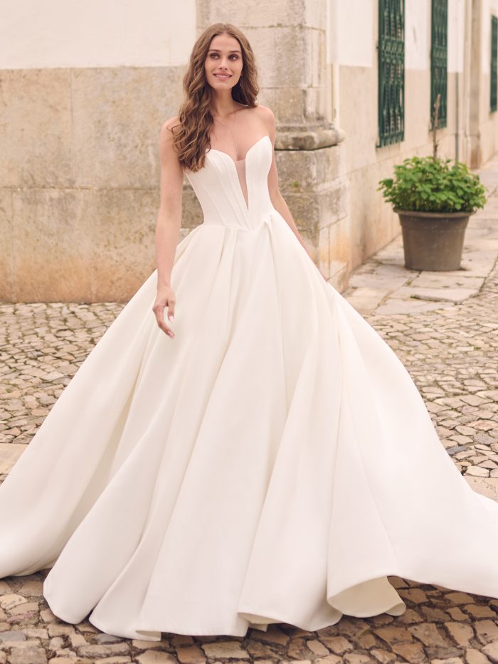 Bride in strapless ball gown wedding dress with deep illusion plunge neckline, structured Mikado bodice, and voluminous pleated skirt, styled on cobblestone streets