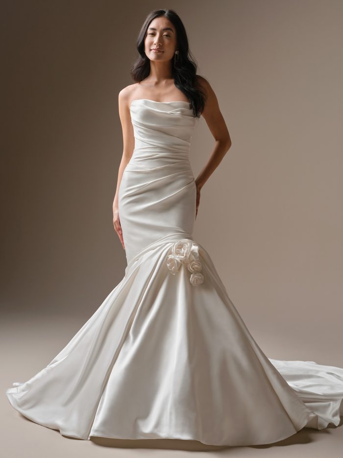 Emelina by Maggie Sottero — strapless satin fit-and-flare wedding dress featuring ruched bodice detailing, draped skirt, and dimensional rosette accents.