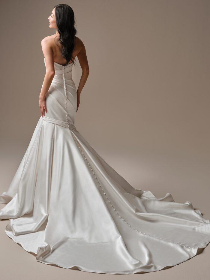 Back view of Emelina by Maggie Sottero — sleek satin fit-and-flare gown with covered buttons trailing down the train and softly ruched detailing at the hips.