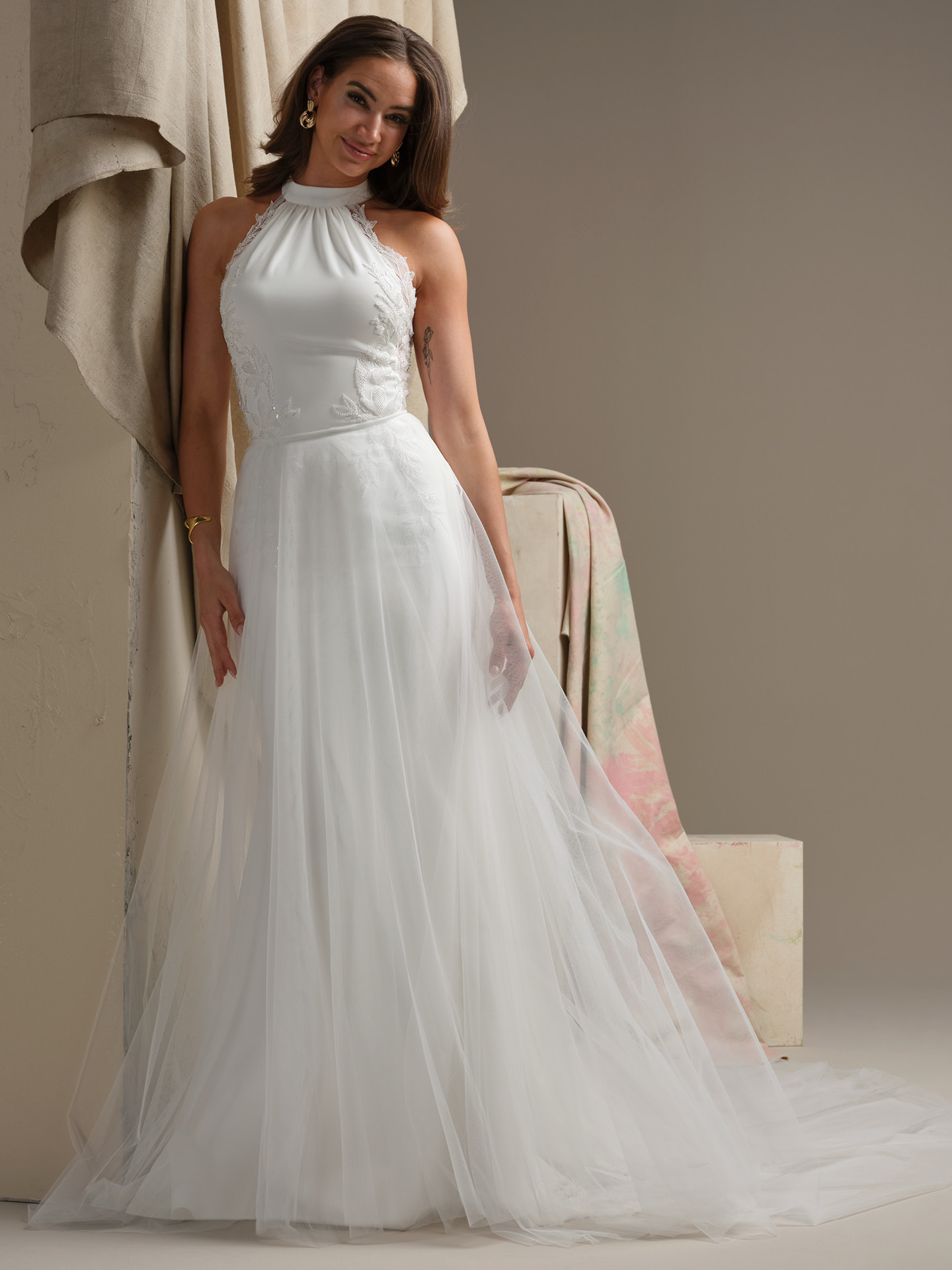 Bride wearing a plain tulle overskirt from the Maggie Sottero Essentials Edit, featuring light, airy layers that add soft volume and a flowing train over a halter-neck lace wedding gown.
