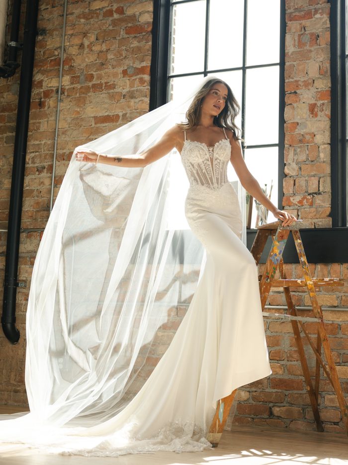 Bride wearing a dramatic beaded lace sheath wedding dress with a sheer corset bodice and thin straps. The gown features intricate embroidery, a curve-hugging silhouette, and a chapel-length train, styled with a long flowing veil for a bold, modern look.