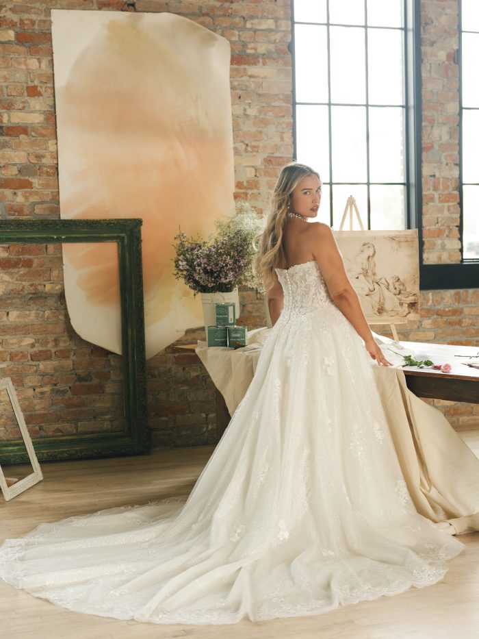 Back view of Jennifer by Maggie Sottero — plus-size lace ballgown wedding gown featuring a chapel-length train, illusion corset bodice, and shimmering floral lace, photographed in soft natural light.