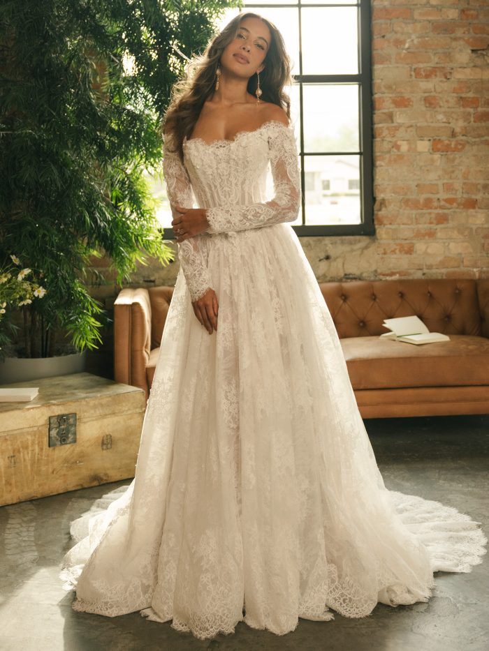 Kitaro wedding dress featuring Chantilly lace, pearl-accented 3D floral appliqués, a strapless off-the-shoulder neckline, long lace sleeves, a basque waistline, and an extended illusion lace train. Model stands in a softly lit studio with greenery and a vintage sofa.
