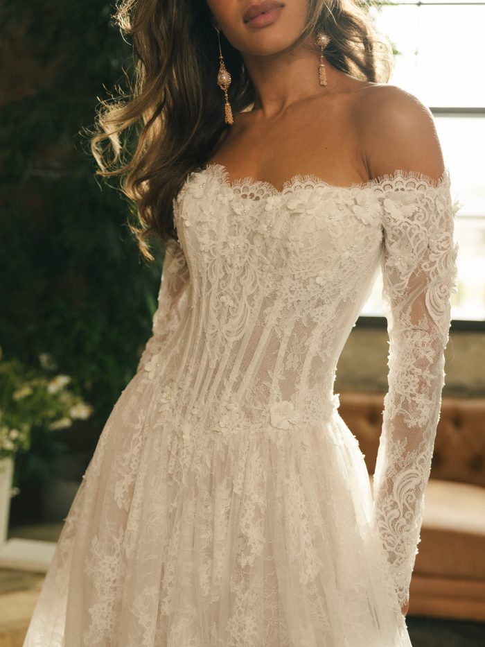 Close-up view of the Kitaro wedding dress showing the scalloped eyelash lace neckline, pearl-accented 3D floral appliqués, exposed boning on the illusion bodice, and detailed long lace sleeves.