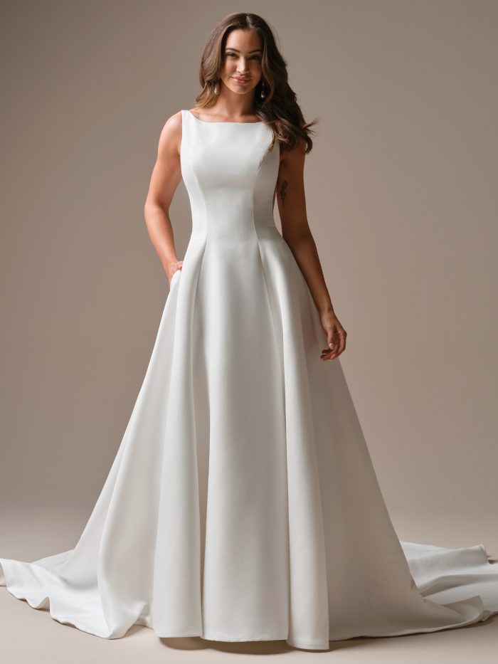 Lawrence by Maggie Sottero — classic bateau-neck satin A-line wedding dress with clean tailoring, pockets, and a flowing train, styled with soft waves in a minimalist studio setting.