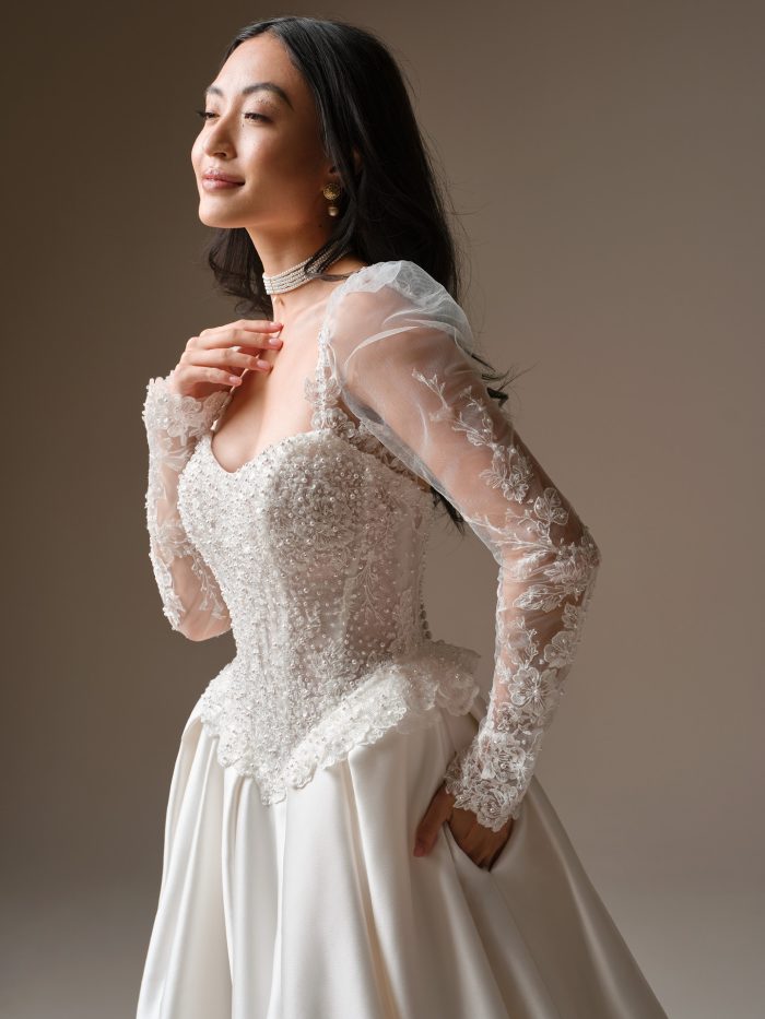 Close-up view of the London wedding dress showing the beaded lace bodice, illusion long sleeves, soft sweetheart neckline, lace tiered peplum detailing, and Cartago satin skirt with pockets.