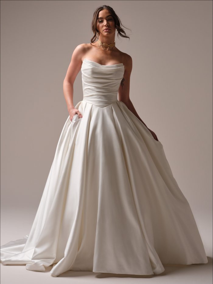 Model wearing the Maureen by Maggie Sottero strapless ballgown wedding dress with a softly draped bodice, structured waistline, full skirt, and pockets in a studio setting.