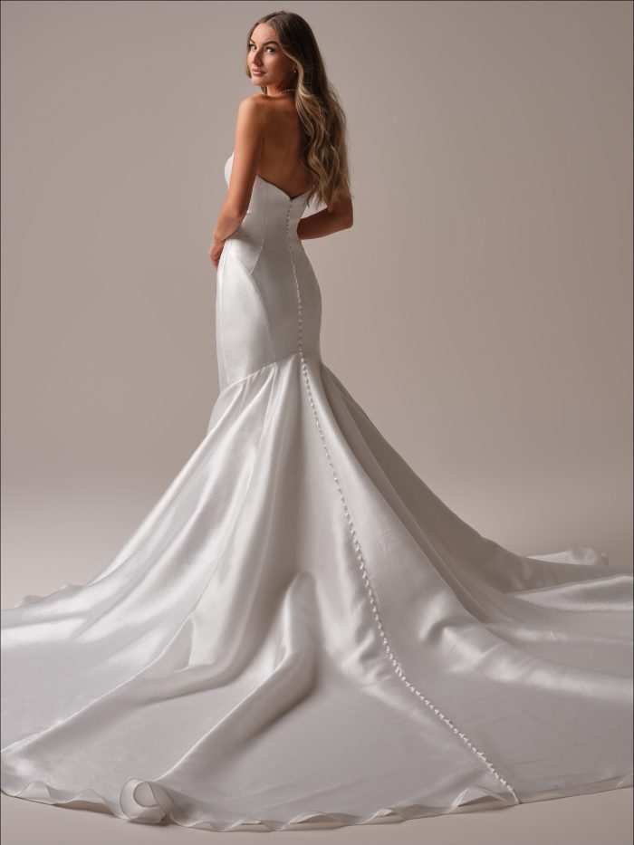 Odette Marie by Maggie Sottero — back view of a strapless satin fit-and-flare wedding dress featuring covered buttons, contour seaming, and a dramatic flowing train, styled with loose waves in a studio setting.