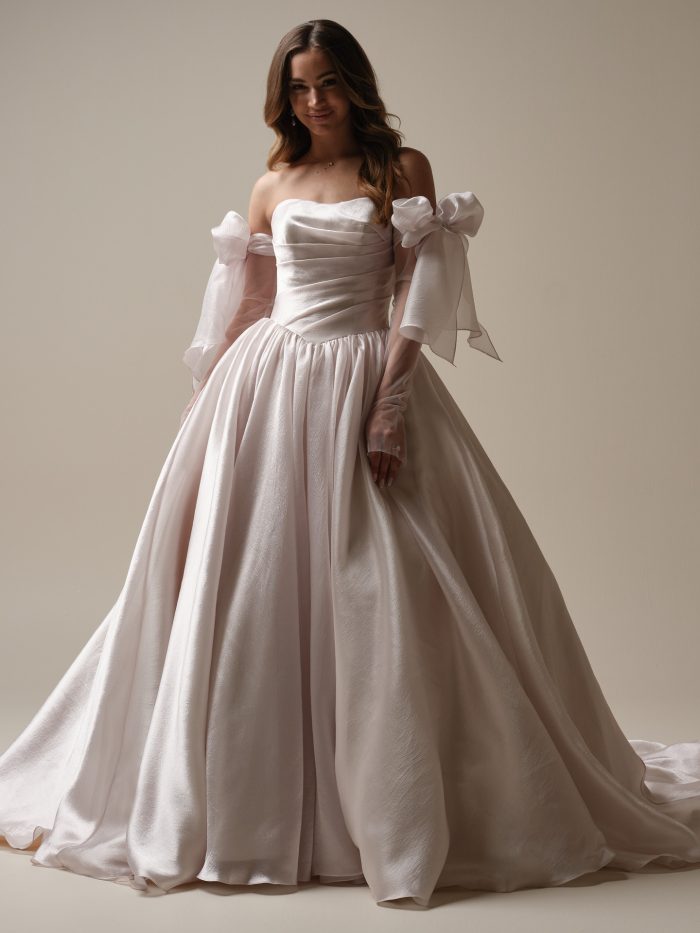 Paris by Maggie Sottero — romantic strapless ballgown wedding dress featuring a ruched bodice, full textured skirt, and detachable voluminous organza sleeves with dramatic bows, styled in a soft neutral studio setting.