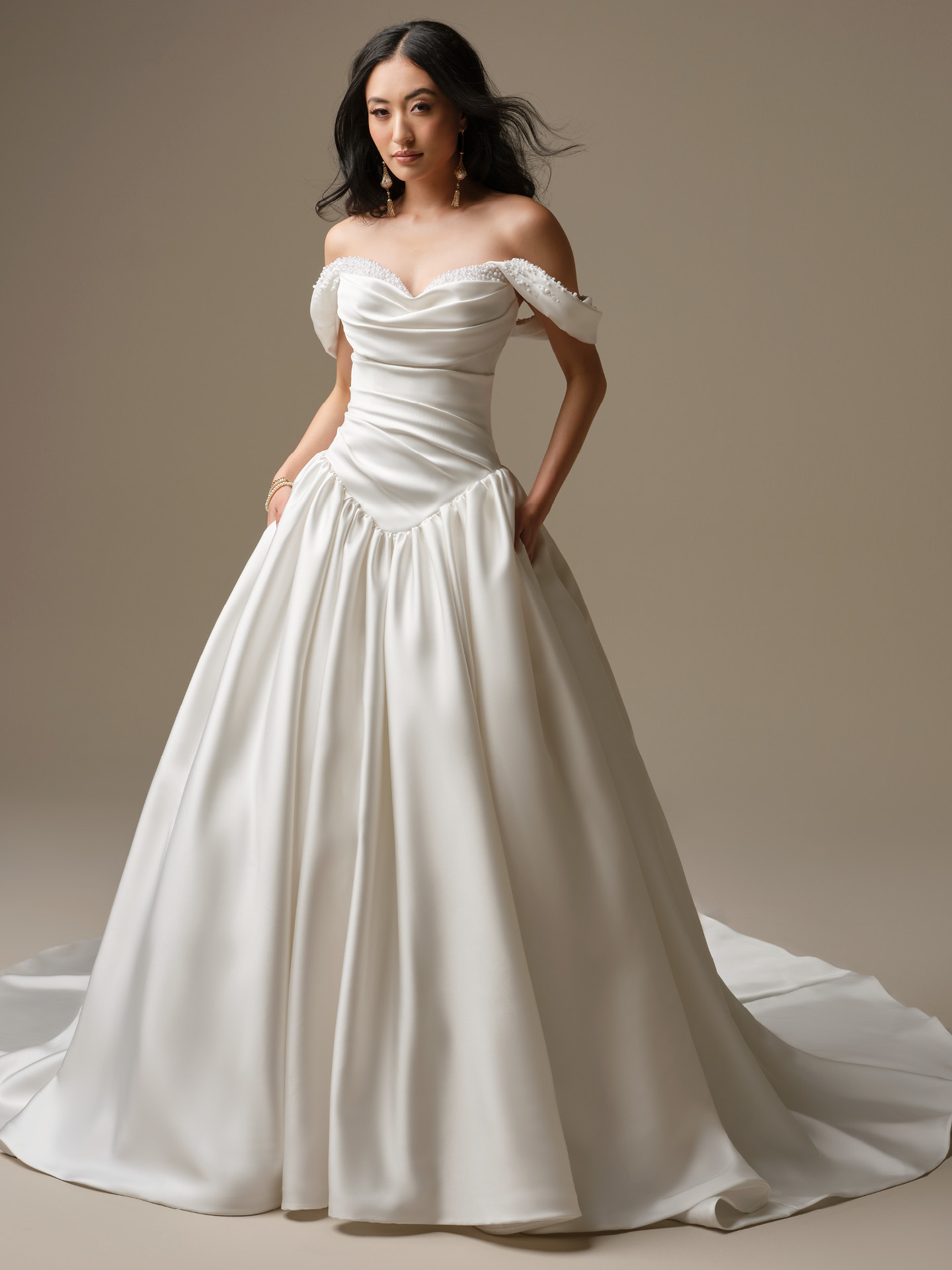 Bride wearing Pasadena by Maggie Sottero, an off-the-shoulder satin ballgown wedding dress featuring a draped sweetheart bodice with pearl-accented straps and a full gathered skirt with a sweeping train.