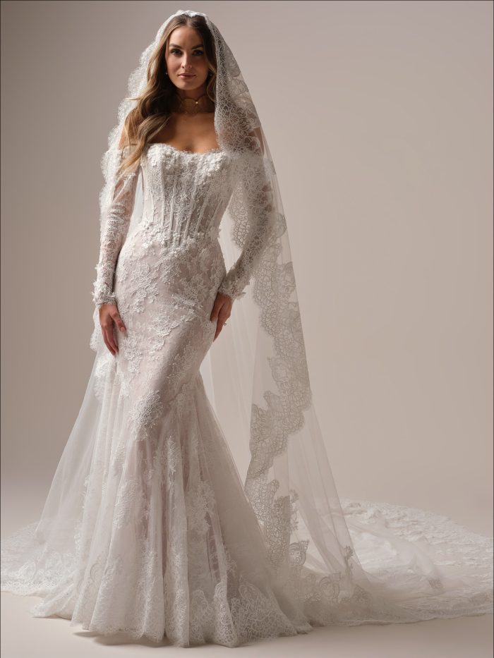 Bride wearing the Maggie Sottero Takera fit-and-flare wedding dress in Chantilly lace with pearl-accented 3-D floral appliqués, strapless off-the-shoulder neckline, long illusion lace sleeves, and extended scalloped lace train. Styled with a dramatic matching lace-edged veil.