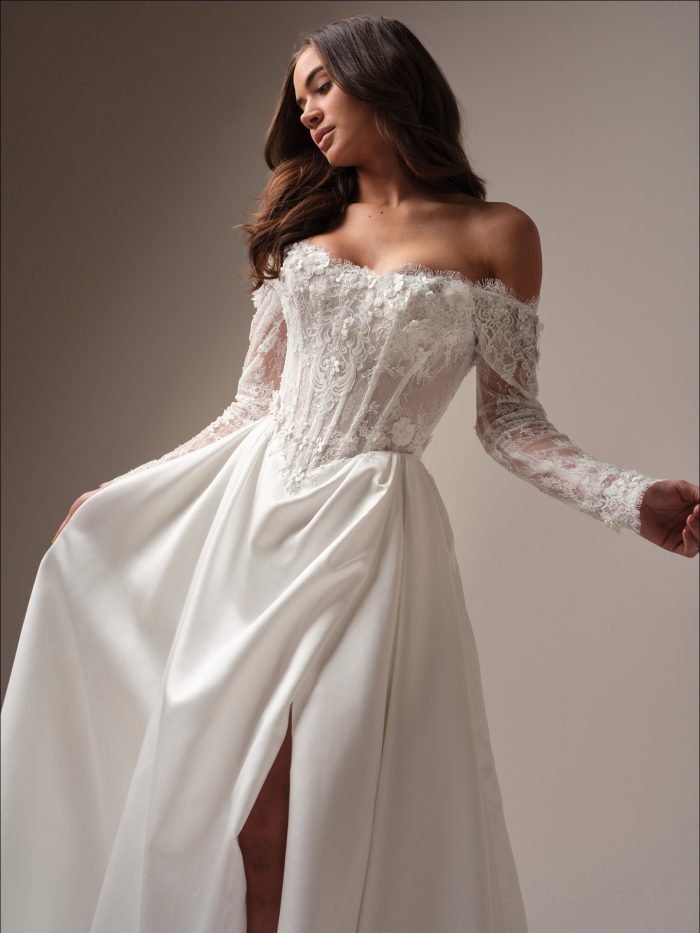 Takera Lane by Maggie Sottero — romantic off-the-shoulder lace A-line wedding dress featuring long illusion lace sleeves, a 3-D floral corset bodice, and a soft satin skirt with a thigh-high slit, styled in a minimal studio setting.