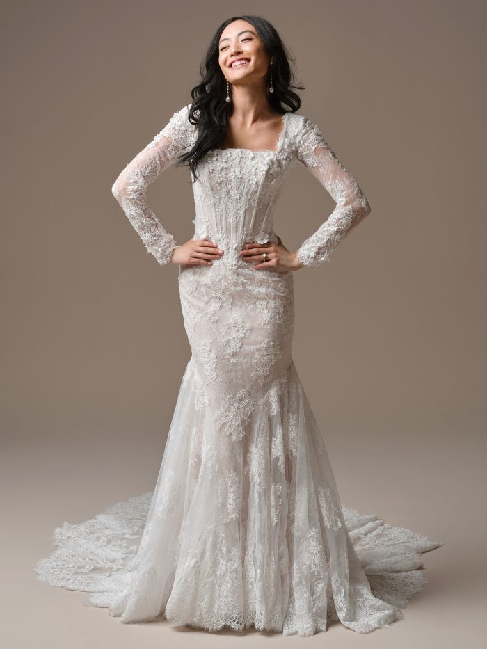Model wearing the Takera Leigh fit-and-flare wedding dress, featuring long illusion lace sleeves, a square neckline, and a sculpted lace bodice that flows into a soft, flared train for a romantic, vintage-inspired look.