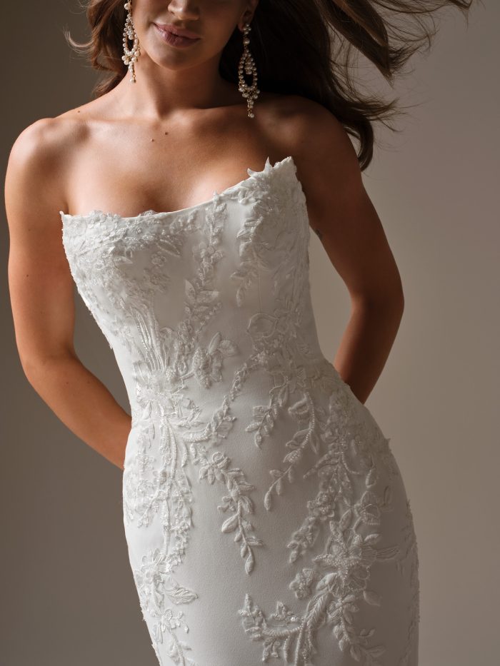 Close-up of a strapless fit-and-flare wedding dress with a sculpted cat-eye neckline and intricate sequin lace embroidery. The bride’s pearl statement earrings complement the gown’s romantic texture and clean, modern silhouette.