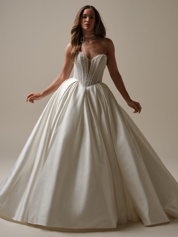 Bride wearing a strapless satin ball gown wedding dress with a pearl-embellished corset bodice and structured sweetheart neckline, standing in soft studio lighting.