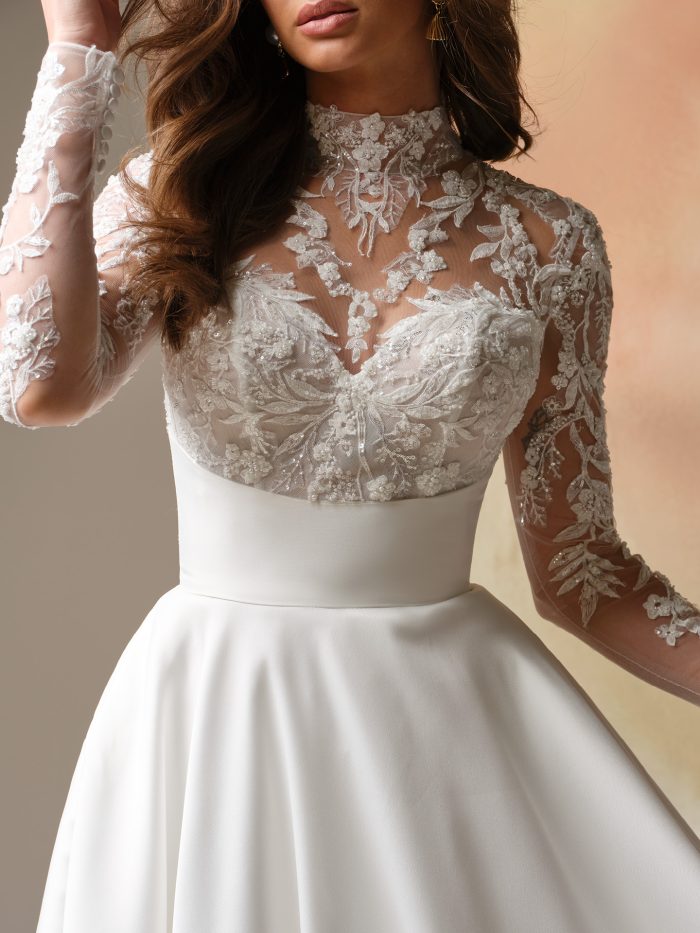 Close-up of Trinity by Maggie Sottero, illusion beaded lace bodice with mock lace turtleneck and long sleeves over classic Mikado skirt, vintage-inspired A-line bridal gown.