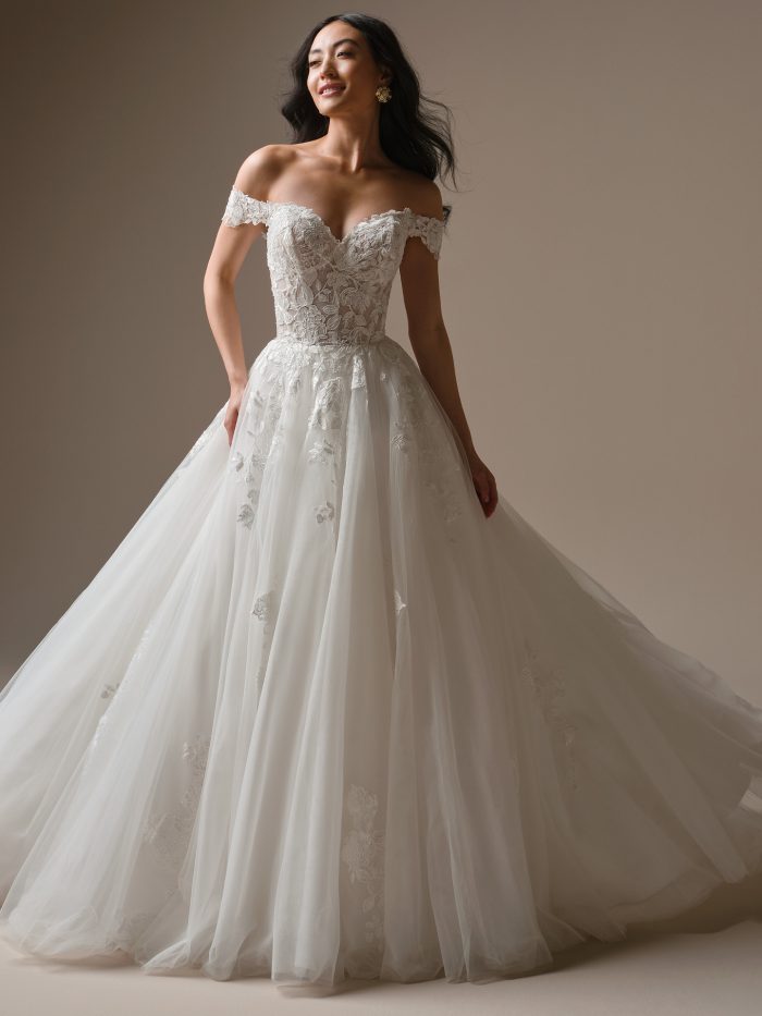 Model wearing the Verina ball gown wedding dress by Maggie Sottero, featuring an off-the-shoulder lace bodice, floral lace appliqués, and a full tulle skirt with soft volume.