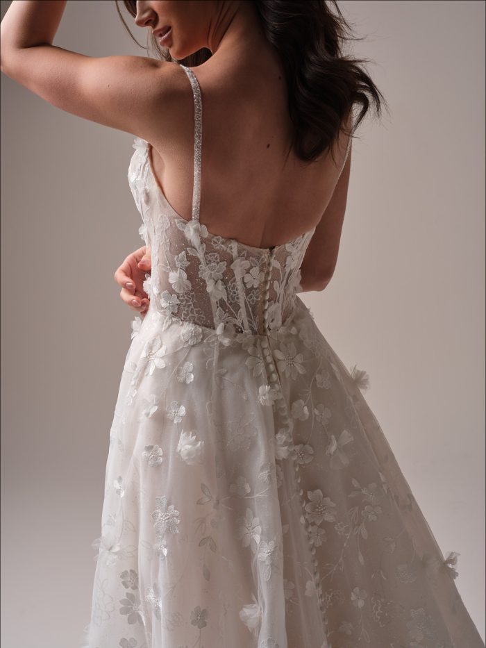 Back view of the Maggie Sottero Wallis ballgown wedding dress showcasing an illusion lace bodice with button detail, pearl-dusted straps, and layered floral motifs flowing into a luxe textured skirt.