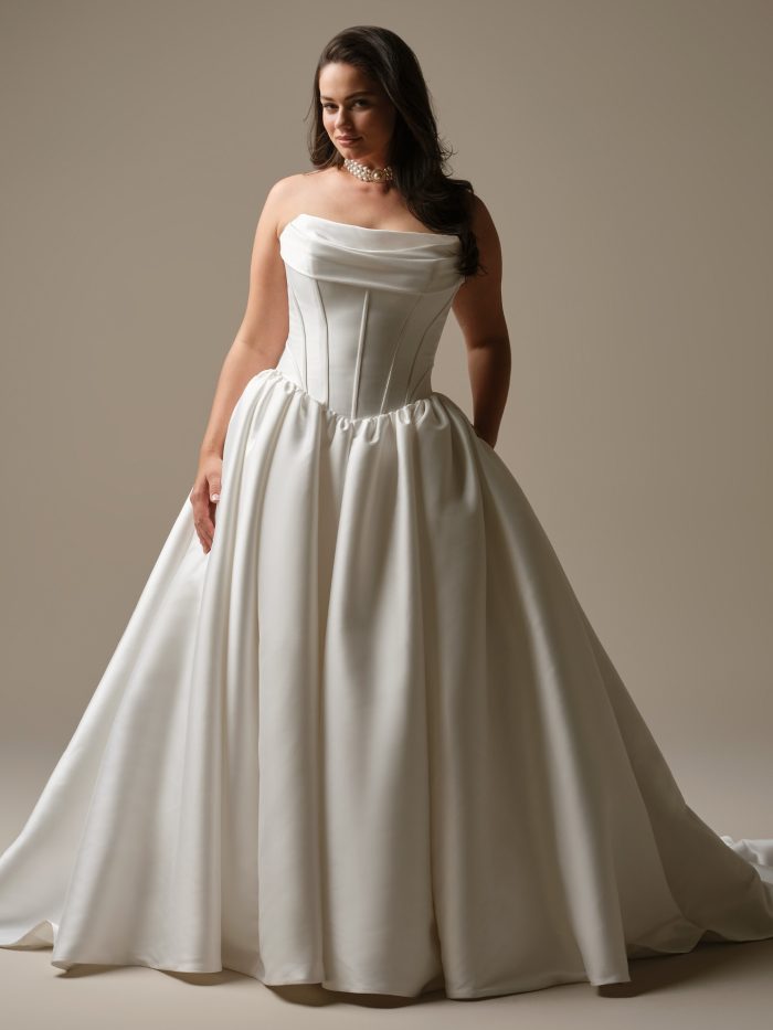 Yuma by Maggie Sottero — strapless satin ballgown wedding dress featuring a draped neckline, structured corset bodice, and full gathered skirt, styled with pearl jewelry in a neutral studio setting.
