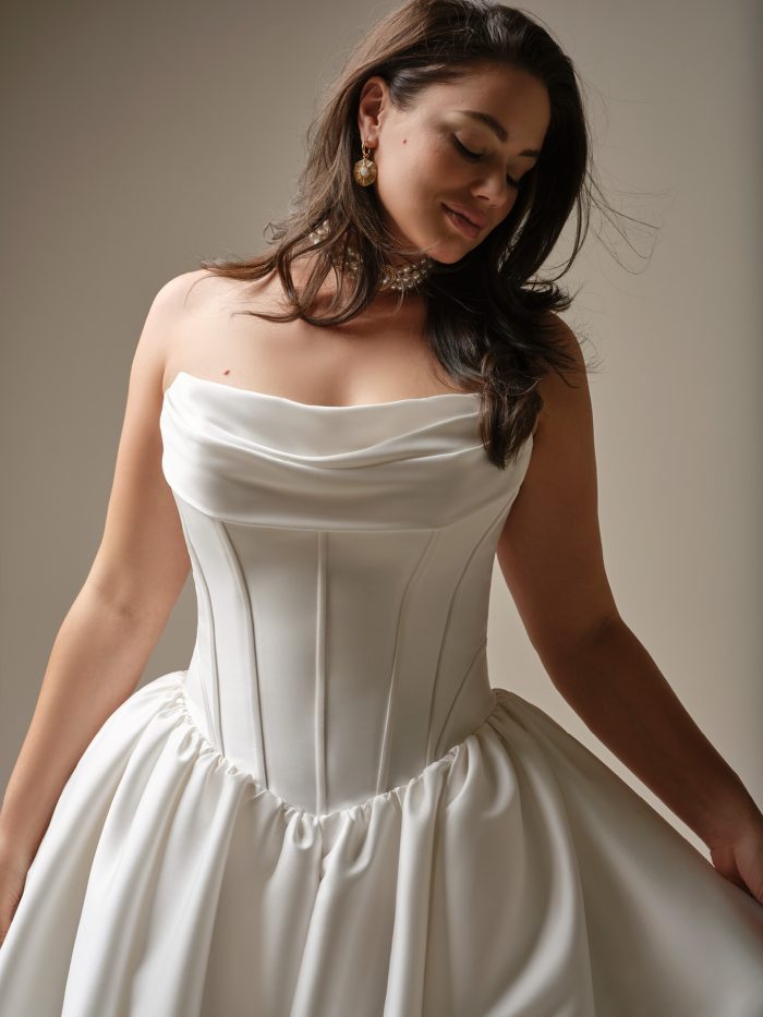 Yuma by Maggie Sottero — close-up of a strapless satin ballgown wedding dress with a draped neckline and structured corset bodice, styled with pearl jewelry and soft waves in a studio setting.