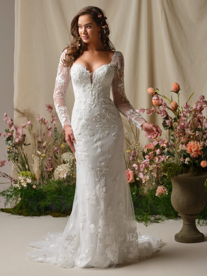 Aubra wedding dress featuring beaded lace motifs over tulle, a lined sweetheart bodice with a plunging illusion neckline, long illusion lace sleeves with fitted shaping, and an extended illusion train accented with lace. Model stands in a floral studio setting.