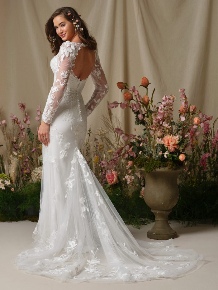 Back view of the Aubra wedding dress showing the keyhole back, long illusion lace sleeves with covered button closures at the wrists, beaded lace motifs over tulle, and an extended illusion lace train flowing behind the model.