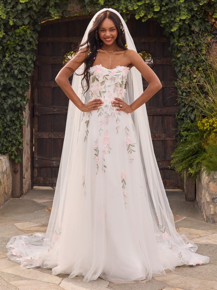 Bride wearing the Blossom A-line wedding dress by Rebecca Ingram, featuring pink 3D floral appliqués, leafy embroidery, and a strapless sweetheart neckline, styled with a long veil in a garden courtyard setting.