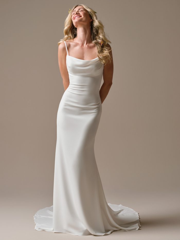 Bride wearing the Brenda gown, a sleek ivory sheath wedding dress with a soft cowl neckline and thin straps, standing in a relaxed, joyful pose with the train flowing behind her.