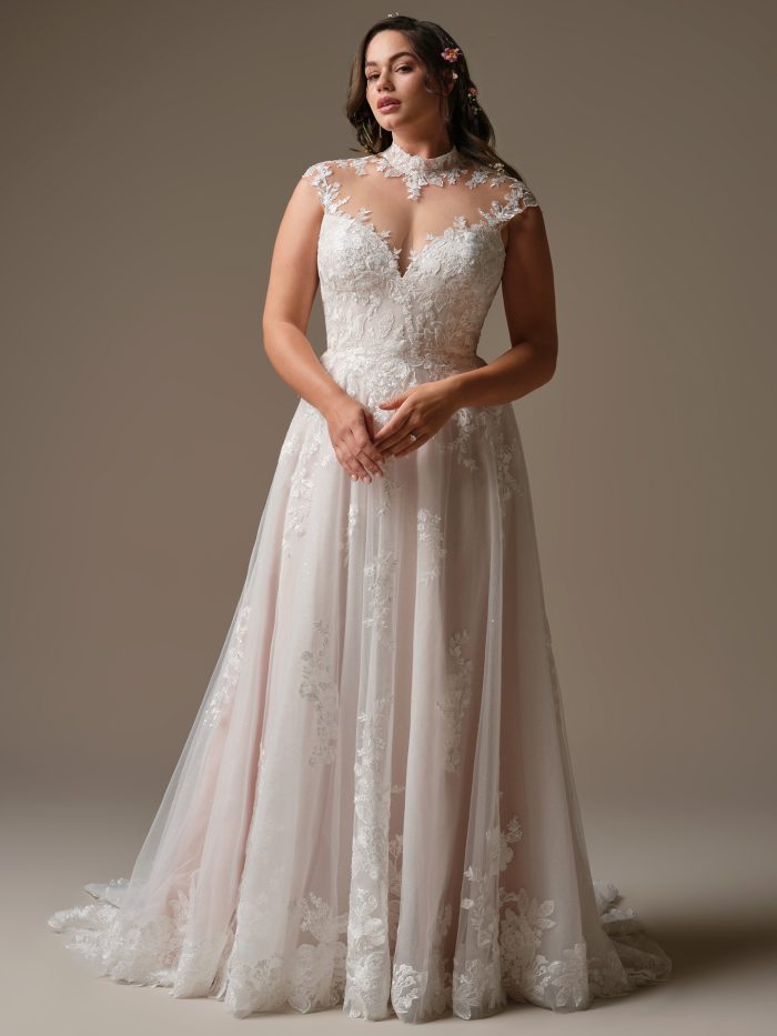 Bride wearing the Rebecca Ingram Cody A-line wedding dress featuring a high illusion lace neckline, cap sleeves, floral appliqué bodice, and soft tulle skirt in a curve size.