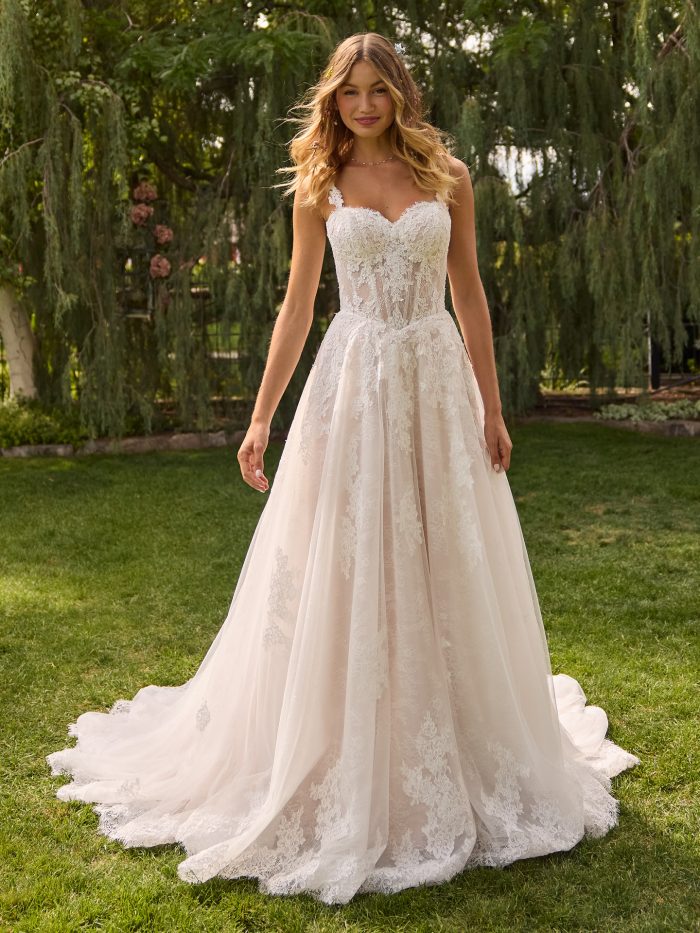 Bride wearing Esmeralda by Rebecca Ingram, an A-line lace wedding dress with a sweetheart neckline, exposed boning, and delicate beaded straps, shown in ivory over blush.