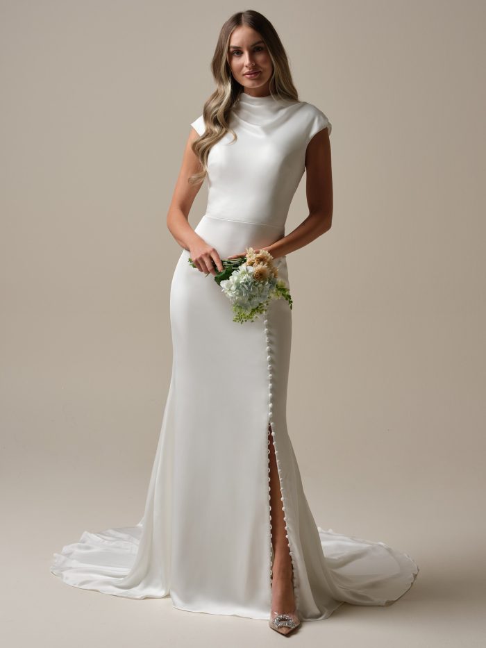 Bride wearing Jett Leigh by Rebecca Ingram, a modest crepe-back satin sheath wedding dress with high cowl neckline, cap sleeves, and covered buttons trailing down the front slit and train.