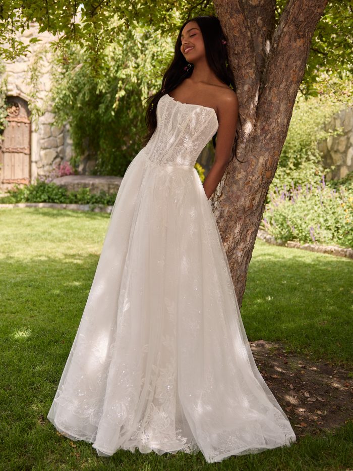 Bride wearing Lisa by Rebecca Ingram, a strapless ball gown wedding dress with a sheer lace bodice, leafy embroidery, and shimmering tulle skirt, shown in ivory over blush.