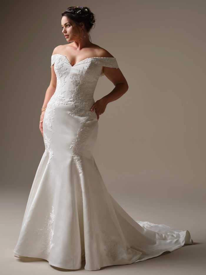 Curve model wearing Miley by Rebecca Ingram — off-the-shoulder fit-and-flare wedding dress with embroidered floral lace, soft satin finish, and chapel-length train, styled with floral hair accents.
