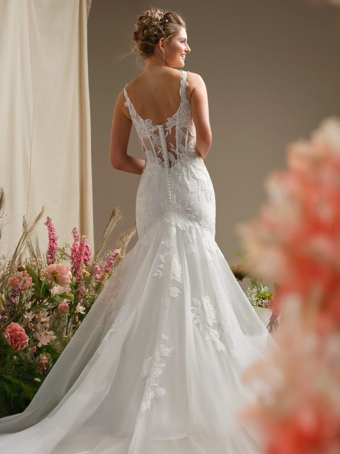 Back view of Pearline by Rebecca Ingram, featuring illusion lace straps, a sheer corset back with covered buttons, and floral lace appliqués trailing down a tulle train.