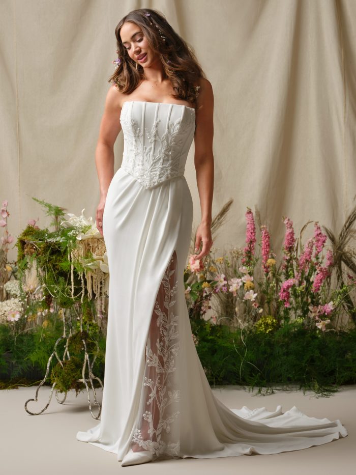 Bride wearing Terry by Rebecca Ingram, a strapless sheath wedding dress with a structured corset bodice, beaded embroidery, and an illusion lace side slit, shown in ivory.