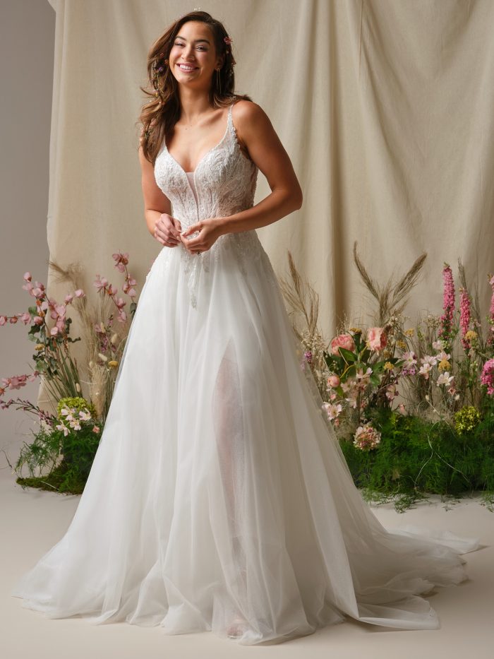 Bride wearing the Valarie A-line wedding dress by Rebecca Ingram, featuring a beaded lace bodice, delicate straps, and flowy tulle skirt with thigh-high slit, styled with floral hair accessories in a garden-inspired setting.