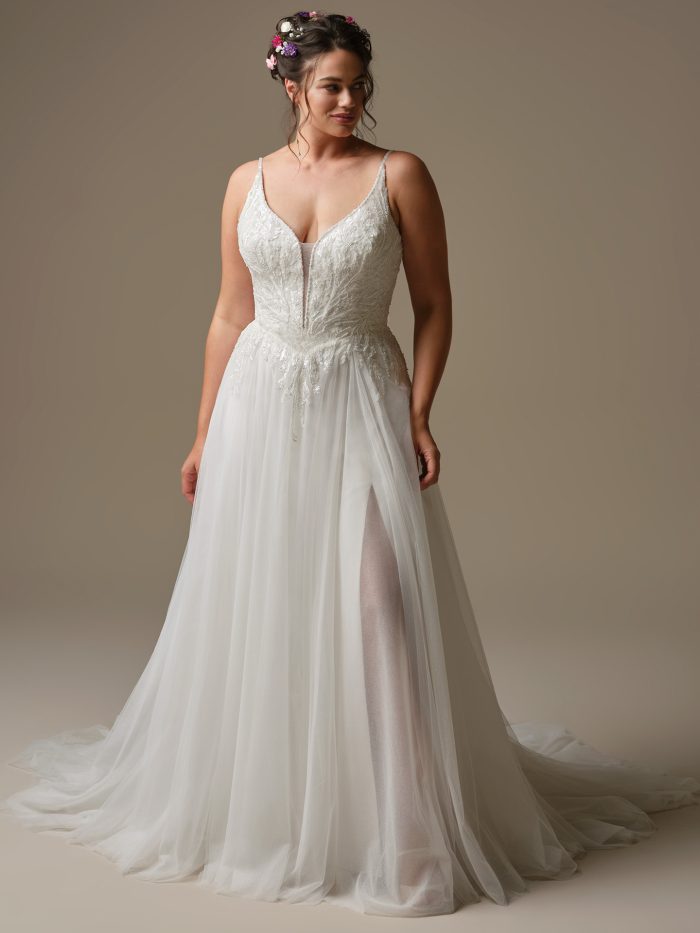 Model wearing Valarie by Rebecca Ingram, an A-line wedding dress with beaded and sequined lace bodice, plunging illusion V-neckline, spaghetti straps, and flowing sparkle tulle skirt with side slit.