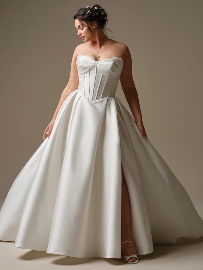 Model wearing the Rebecca Ingram Zorina ballgown wedding dress with a sculpted basque waist, exposed boning, lace-trimmed side slit, and full Mikado skirt.