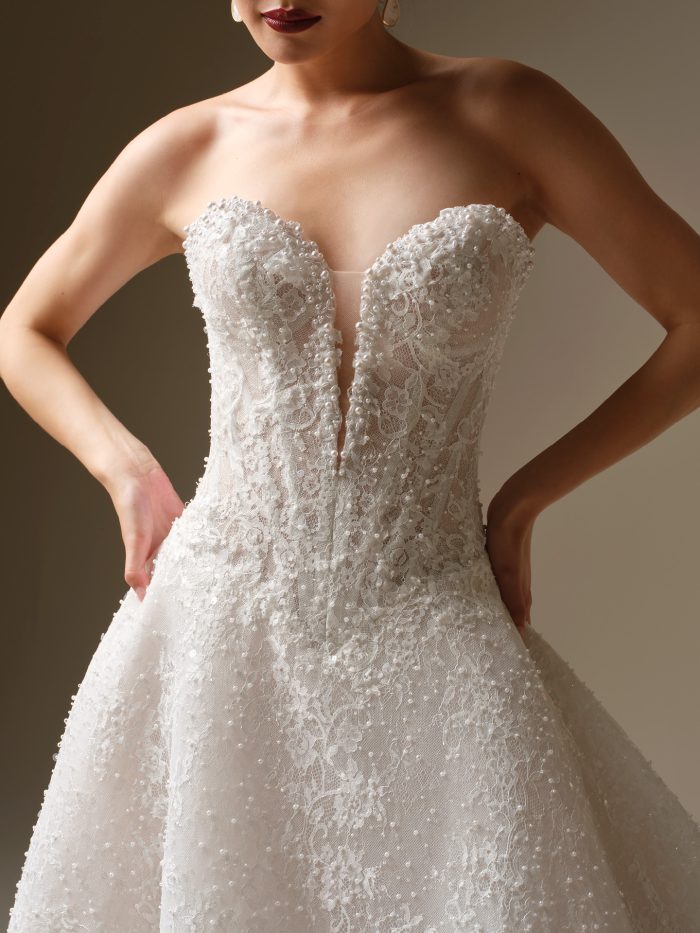Angelica A-line wedding dress with a strapless beaded lace bodice, deep illusion neckline, and intricately textured lace details shown in a close up studio view.