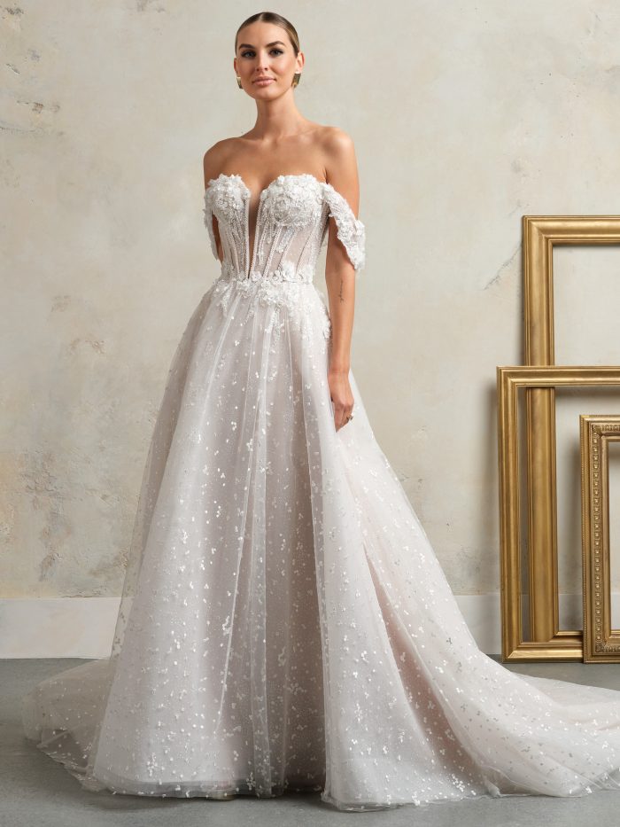 Bride wearing off-the-shoulder A-line wedding dress with illusion plunge neckline, corset bodice adorned with 3D floral appliqués, and glitter tulle skirt with chapel-length train
