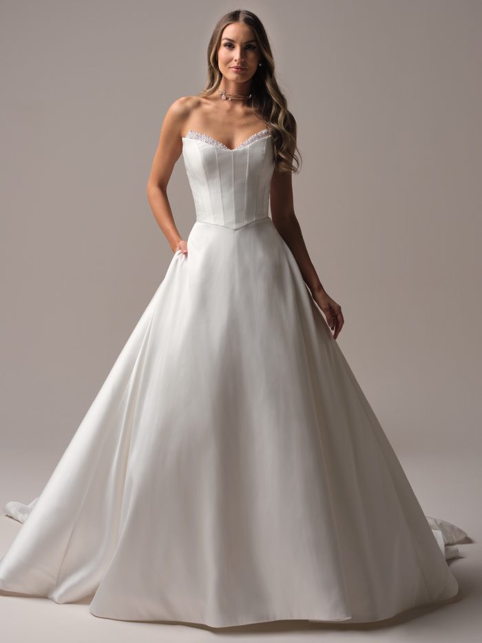 Bride wearing a strapless satin A-line wedding dress with a beaded neckline and structured corset-style bodice, posing gracefully in soft studio lighting.