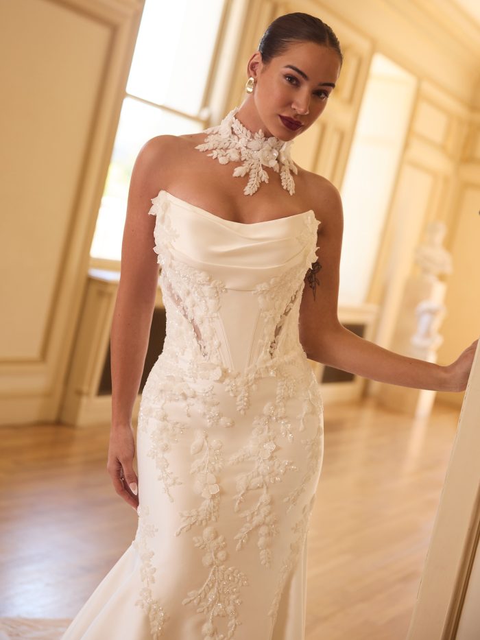 Close-up of Burke by Sottero and Midgley, a strapless fit-and-flare wedding dress with a draped satin neckline, exposed corset bodice, and beaded floral lace appliqués, styled with a matching lace choker.