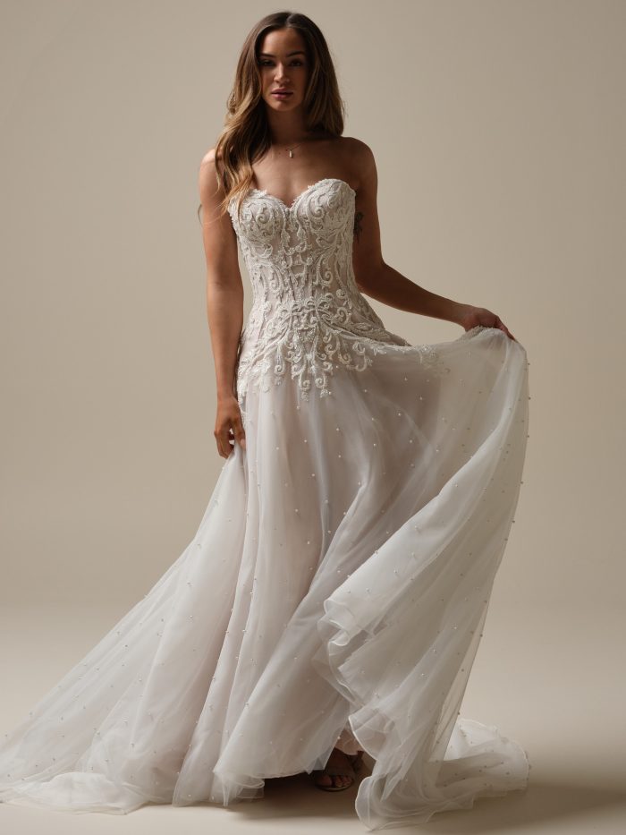 Bride wearing the Dharma A-line wedding dress with a strapless sweetheart neckline, scattered pearl tulle skirt, and ornate lace bodice, holding the skirt to show movement and shimmer.