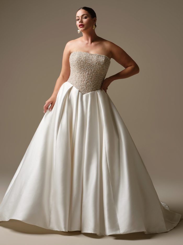 Curve model wearing Dmara by Sottero and Midgley — strapless ballgown wedding dress with fully beaded bodice, satin skirt, and subtle basque waist, styled with statement earrings and sleek updo.