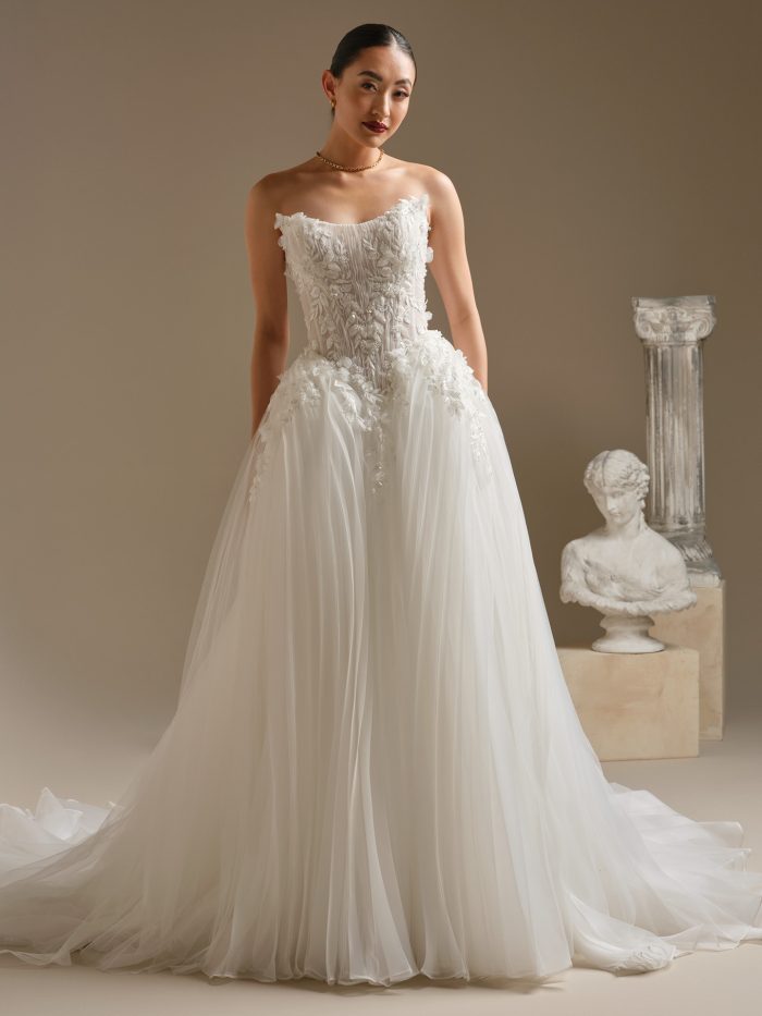 Bride wearing the Jayna ball gown wedding dress by Sottero and Midgley, featuring a strapless cat-eye neckline, illusion bodice with 3-D floral beaded lace, and a full pleated tulle skirt styled in a modern studio setting.