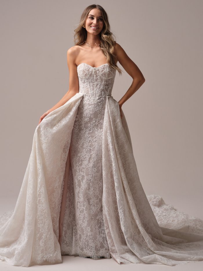 Bride wearing the Jovienne wedding dress with the detachable lace train, showing the strapless sweetheart bodice and dramatic layered lace overskirt for added volume.