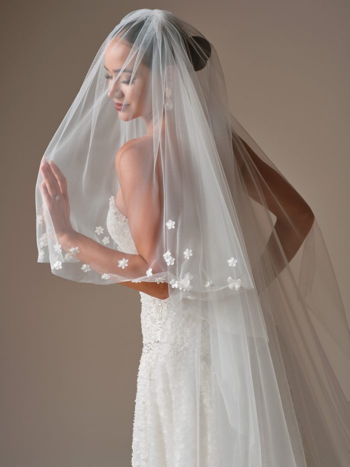 Bride wearing the Marceline veil by Sottero and Midgley, featuring a flowing layered design with scattered 3D floral appliqués along the edges.